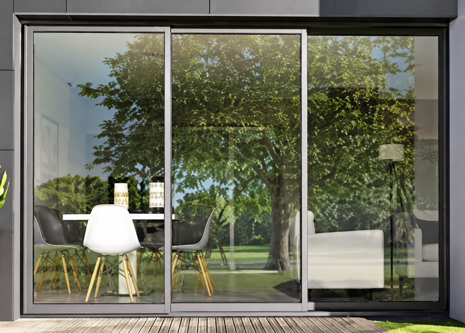 Une grande baie vitrée coulissante à cadre noir, dans laquelle se reflètent des arbres, laisse voir une table et des chaises blanches à l'intérieur.