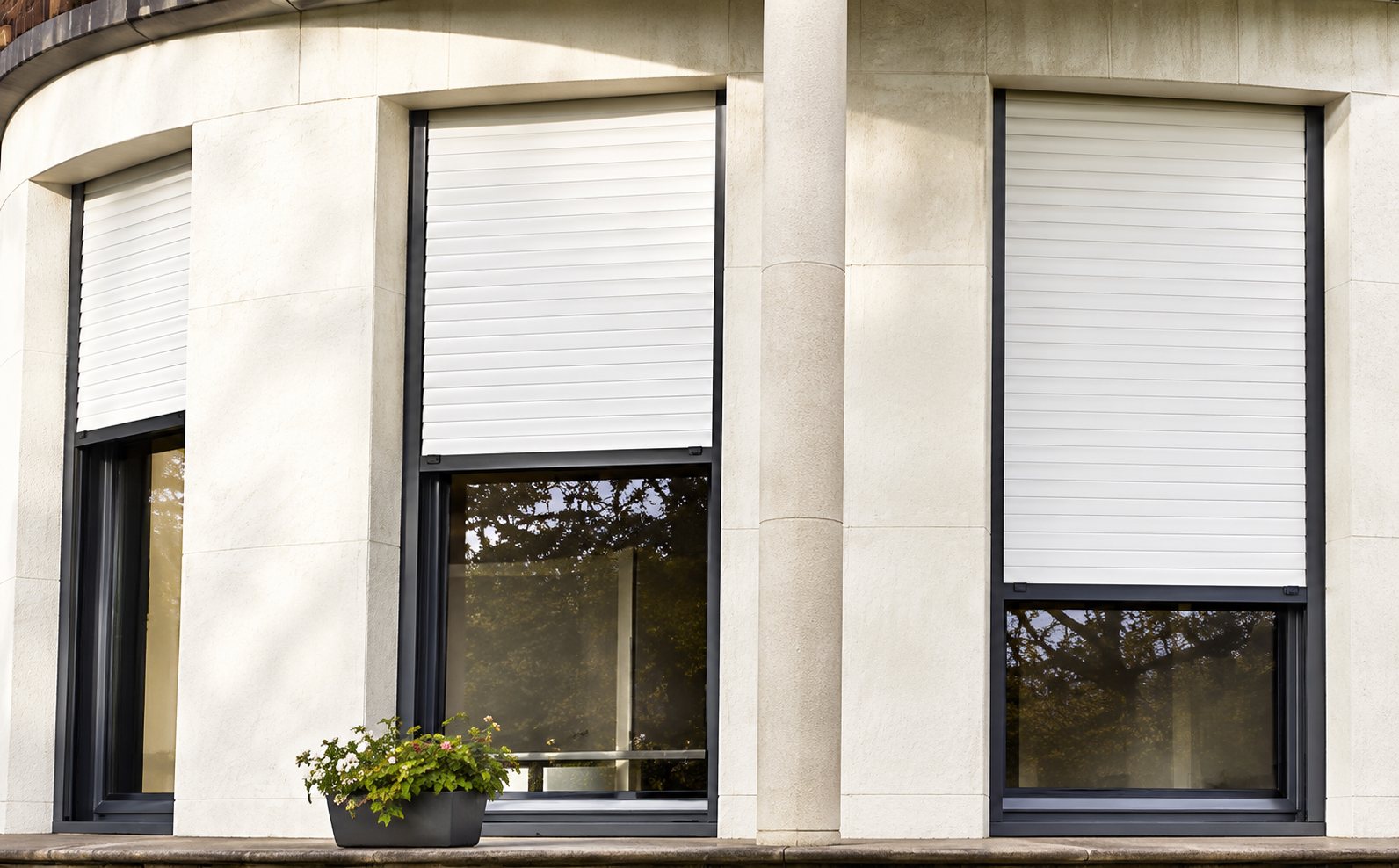Façade d'un bâtiment blanc avec trois fenêtres équipées de volets roulants blancs partiellement baissés, et une petite plante sur un rebord.