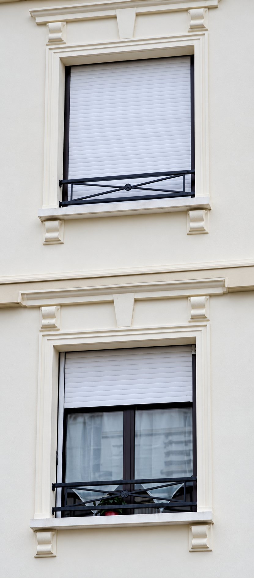 Façade claire d'un immeuble avec deux fenêtres encadrées de moulures, équipées de volets roulants partiellement baissés et de garde-corps métalliques noirs.
