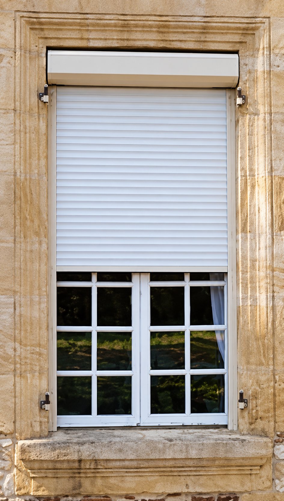Fenêtre blanche à petits carreaux encadrée de pierre de taille, équipée d'un volet roulant blanc à moitié descendu.