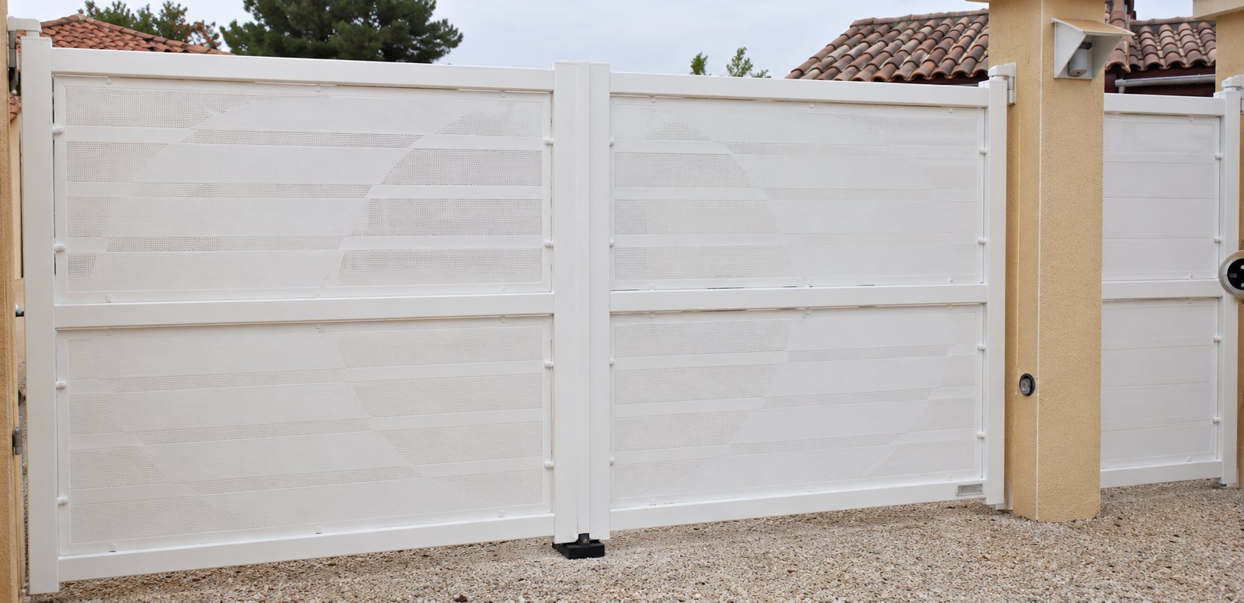 Un grand portail blanc à deux vantaux fermé, encadré par des piliers beiges, devant une maison au toit de tuiles.