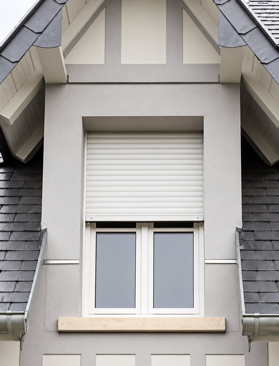 Lucarne d'une maison à toit d'ardoise avec une fenêtre blanche surmontée d'un volet roulant en partie baissé.