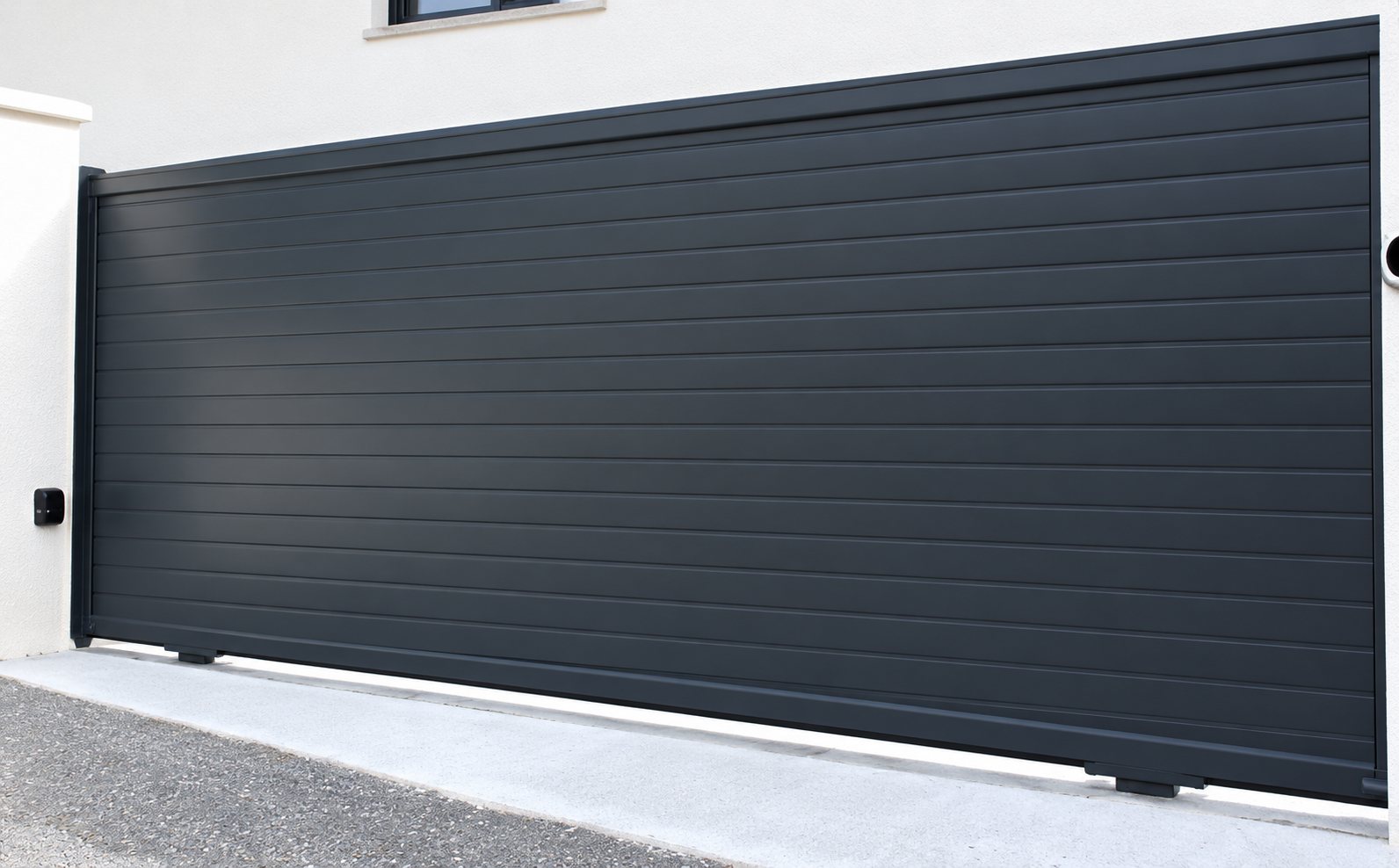 Un grand portail coulissant gris anthracite à lames horizontales installé devant un mur blanc, avec un petit boîtier sur le pilier de gauche.