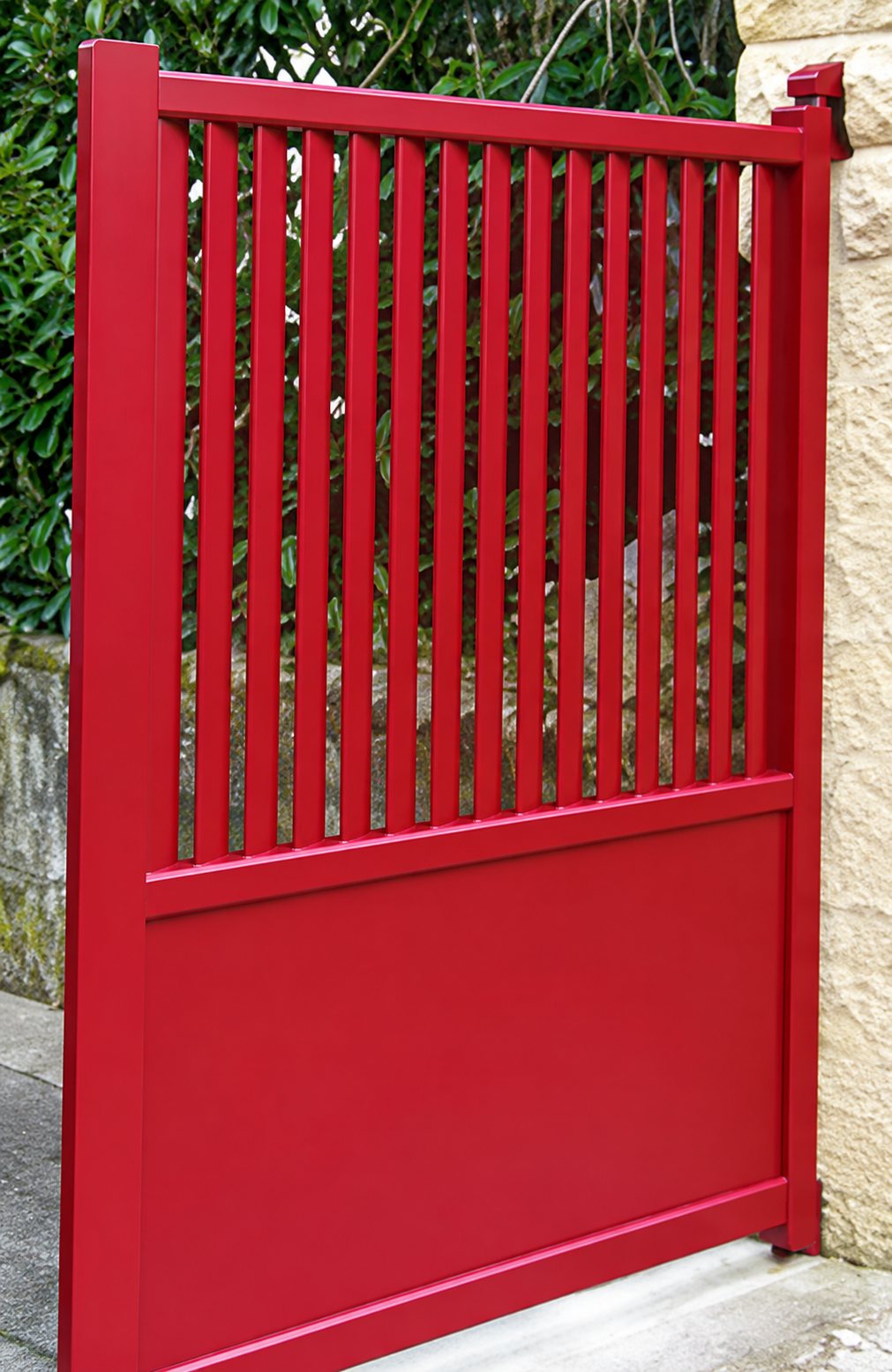Un portail métallique rouge à barreaux verticaux dans la partie haute et panneau plein en bas, fixé à un pilier en pierre claire.