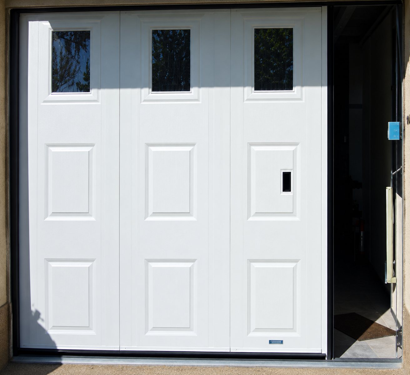 Une porte de garage blanche à panneaux moulés, équipée de trois petits hublots en partie haute, installée dans une façade.