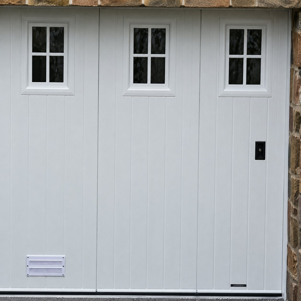 Une porte de garage blanche à lames verticales, équipée de trois petites fenêtres en partie haute et d'une grille d'aération en bas à gauche.