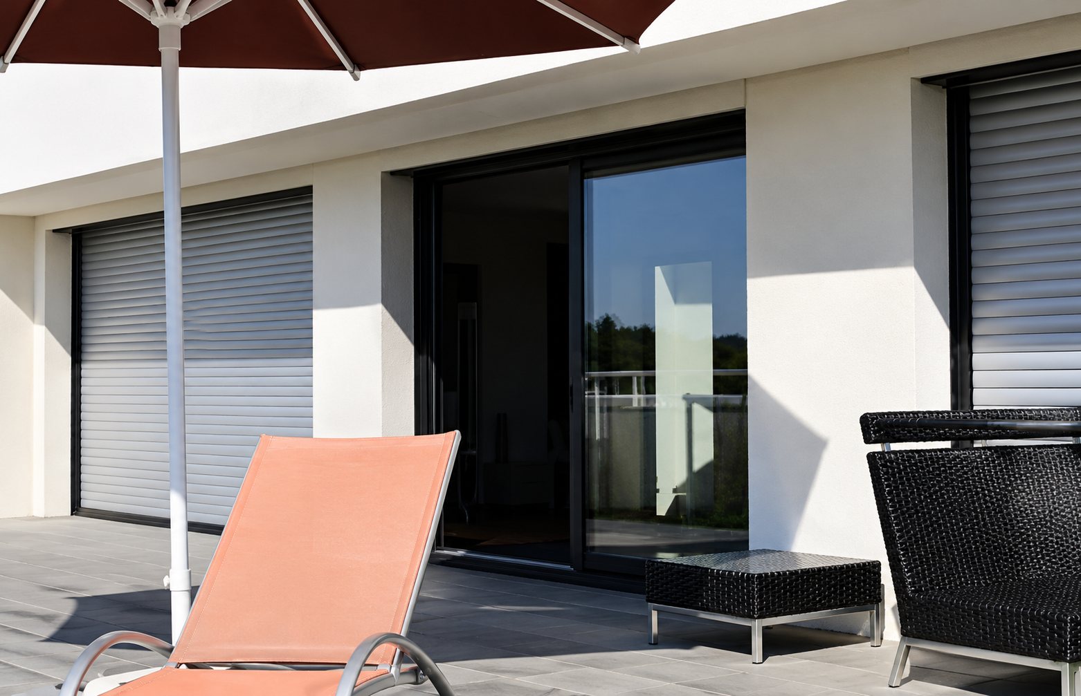 Terrasse en bois d'une maison contemporaine avec un transat orange, un fauteuil noir, un parasol bordeaux et de grandes baies vitrées.