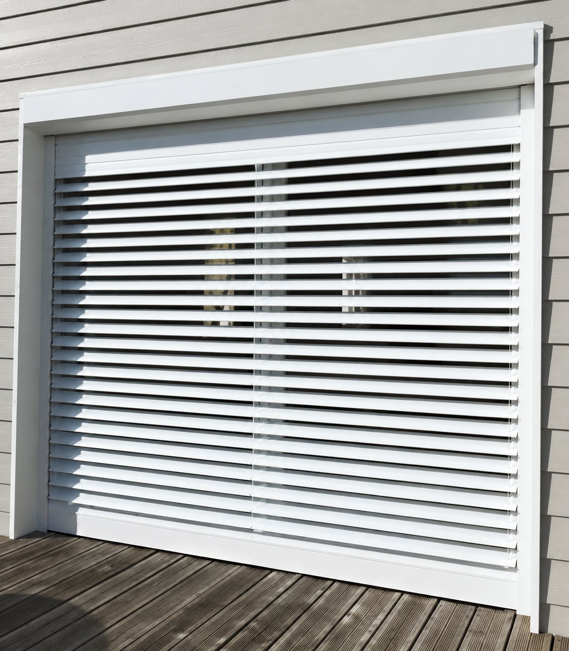 Un grand volet blanc à lames horizontales orientables est installé devant une baie vitrée, au-dessus d'une terrasse en bois.